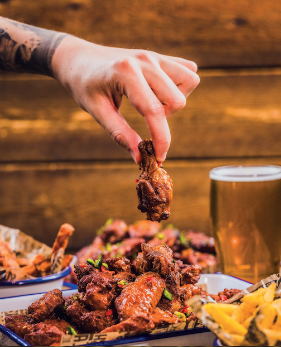 A hand takes a chicken wing from a pile of chicken wings on a plate, with chips and a pint of beer on the side.