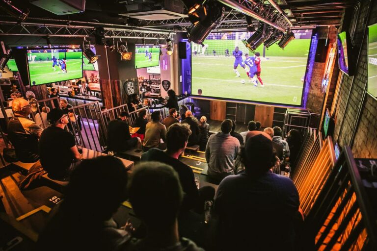 An interior view from the top of the indoor stadium seating at Belushi's bar in Hammersmith, with view of a large projector and smaller HD screens around